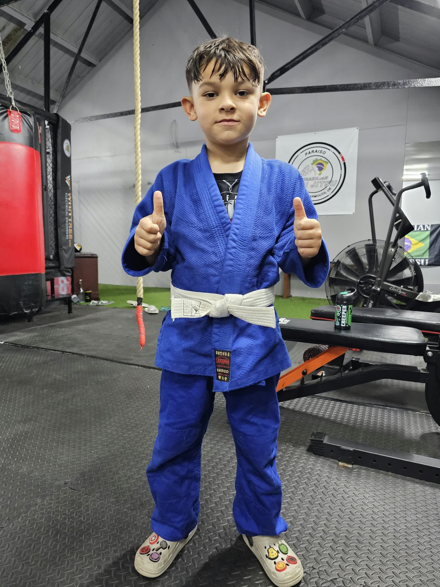 Young Union Thai student in a blue BJJ gi with a white belt giving two thumbs up