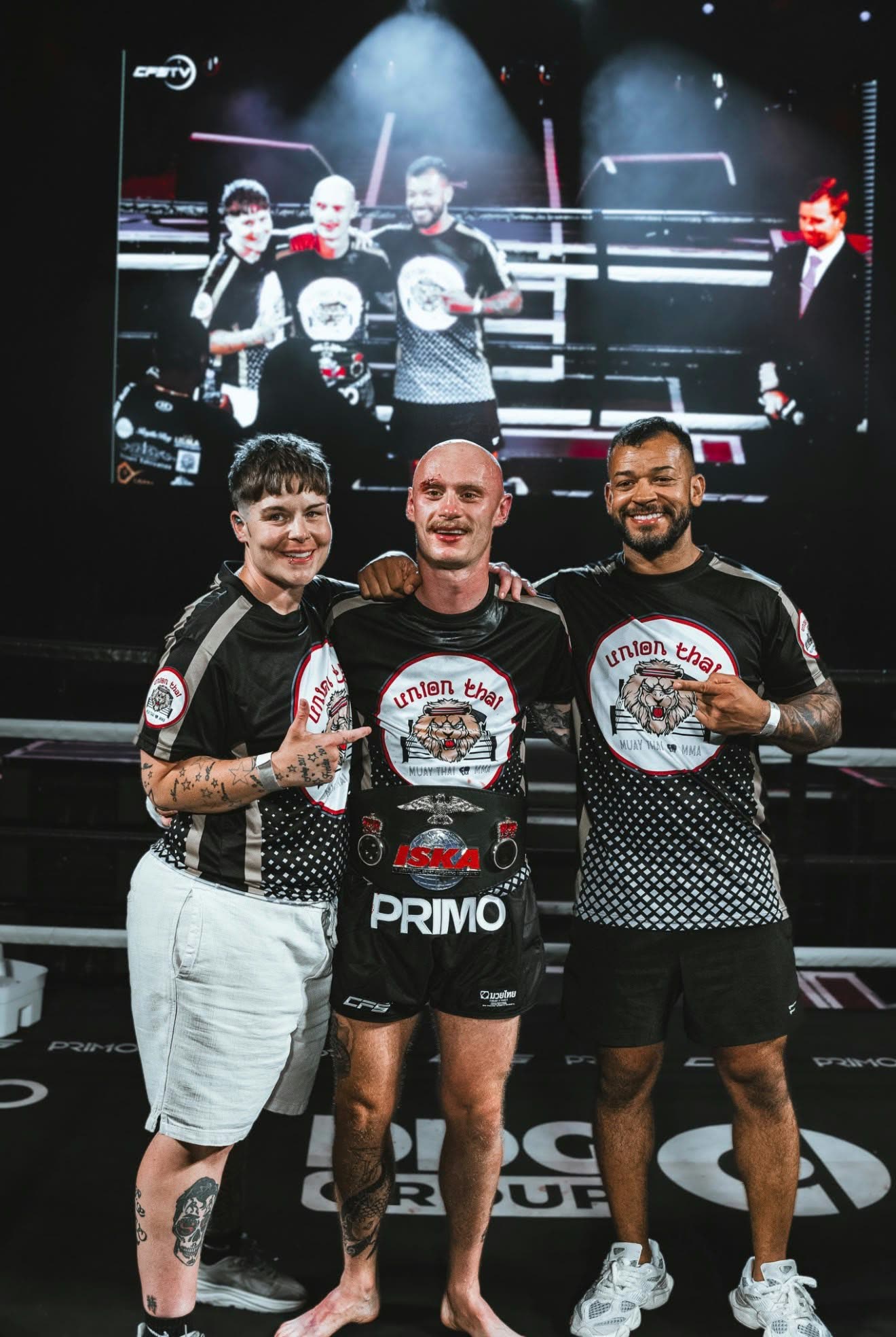 Three Union Thai coaches and fighters posing in the ring after a fight, with the event big screen behind them