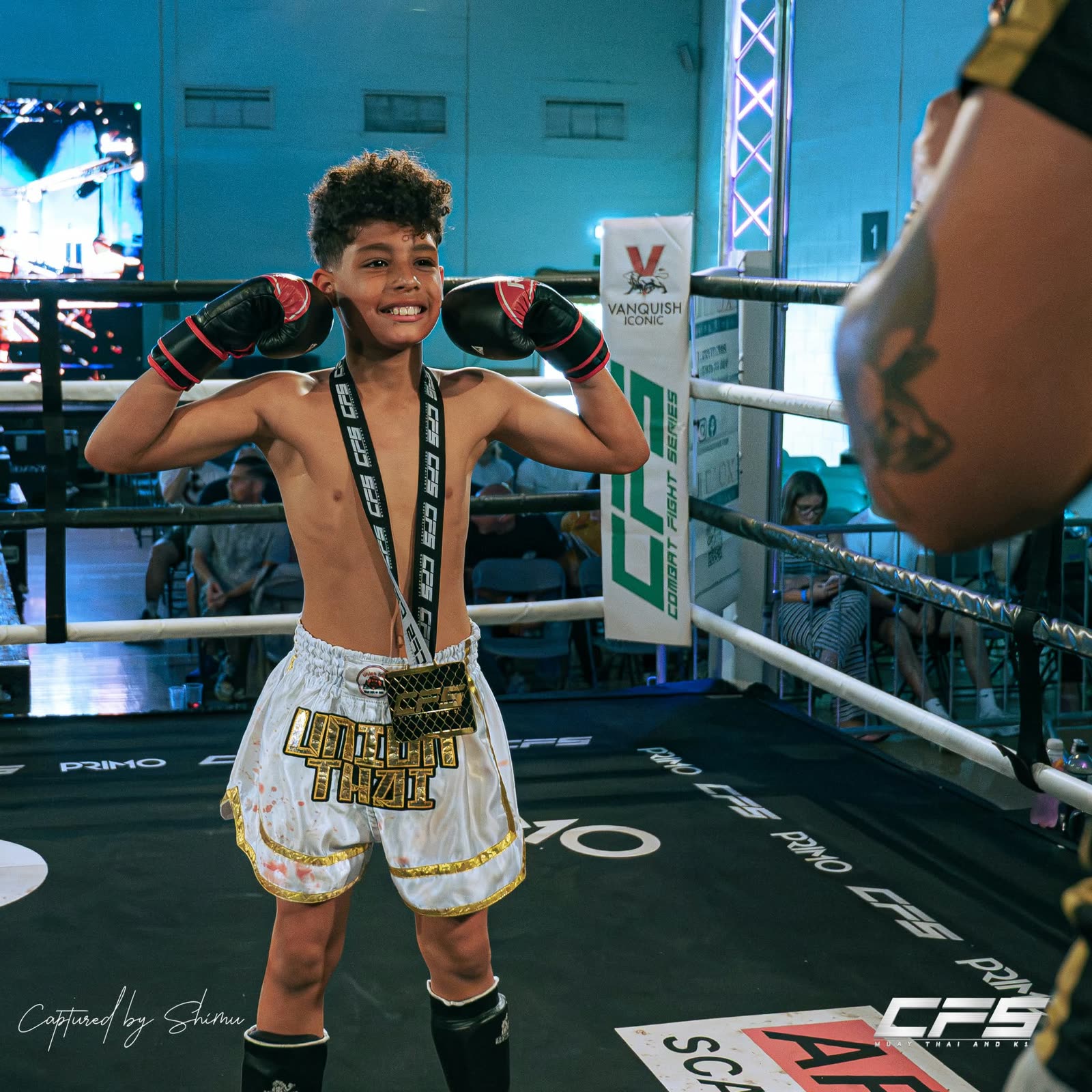 Young Union Thai fighter in white Union Thai shorts flexing in the ring with a medal